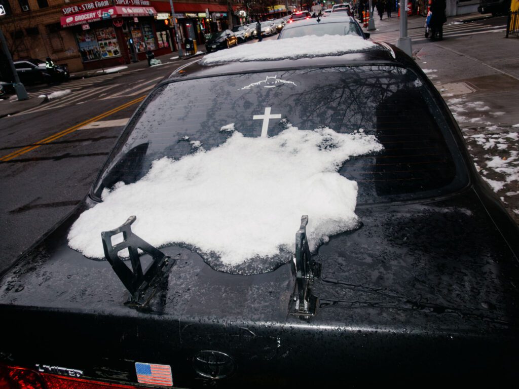 Black and white street photograph of a car's snow-covered rear windshield, with a small Batman symbol sticker and a hanging cross ornament inside it, parked alongside a city sidewalk. In the background, city storefronts and a sidewalk can be seen.
