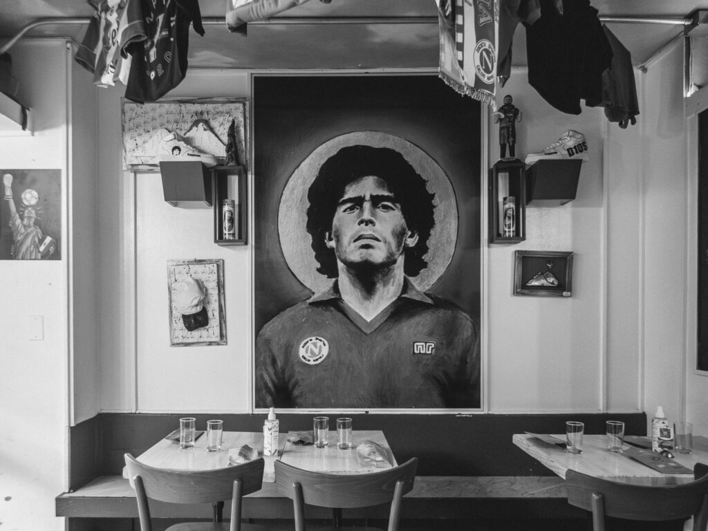 Black and white interior photograph of the restaurant 'Song e Napul' located in NYC. A prominent painting of Diego Maradona wearing a Napoli football jersey is displayed on the wall. Surrounding the painting are various memorabilia, including football jerseys hanging from the ceiling, framed photographs, and a figurine in a showcase. Below, a set of wooden tables and chairs are arranged, with glasses and a menu left on one of the tables.
