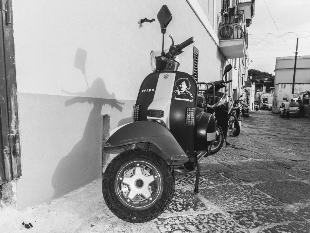 Black and white photo of a narrow Italian street lined with parked Vespas. The closest Vespa features a sticker with the face of Diego Maradona on its side. Its shadow, cast on the adjacent wall, strikingly captures the silhouette of the Vespa's mirrors and handlebars. Traditional buildings with balconies extend down the alley, and the cobblestone road shows signs of wear and age.