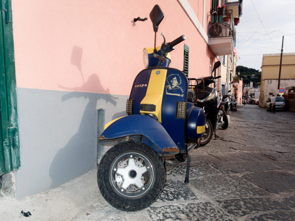 Black and white photo of a narrow Italian street lined with parked Vespas. The closest Vespa features a sticker with the face of Diego Maradona on its side. Its shadow, cast on the adjacent wall, strikingly captures the silhouette of the Vespa's mirrors and handlebars. Traditional buildings with balconies extend down the alley, and the cobblestone road shows signs of wear and age.