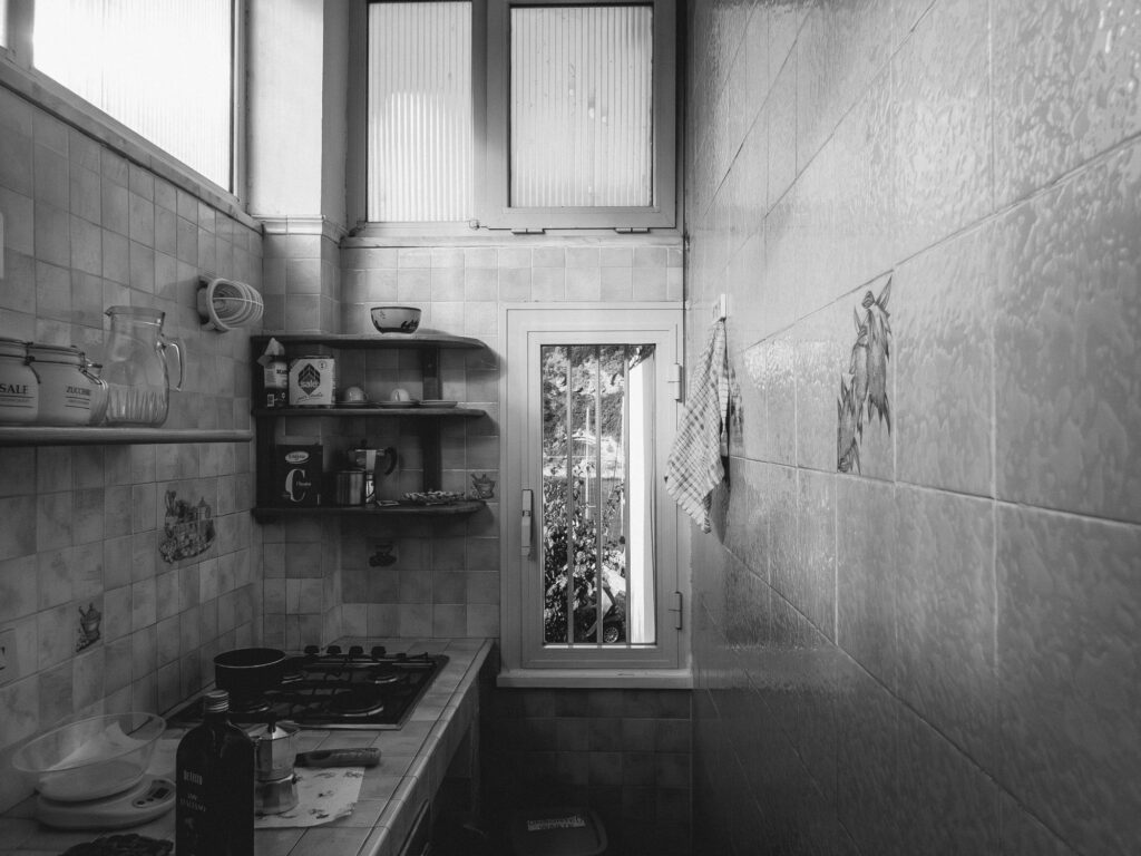 Black and white photograph of a vintage kitchen interior. Ceramic tiles cover the walls, with decorative motifs of vegetables. A wooden shelf holds various kitchen items including a pitcher, bowls, and containers. Below, a gas stovetop with a pan sits next to a countertop with a bottle of olive oil and other cooking essentials. A checkered cloth hangs on the wall near a window that offers a glimpse of the outdoors.