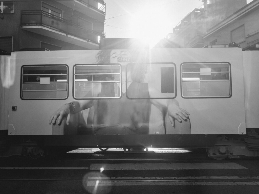 Black and white photograph capturing a city tram with a large advertisement of a woman's face and outstretched hands on its side. The sun beams brightly in the background, casting a flare onto the camera lens. Urban buildings with balconies can be seen in the backdrop, and the tram's undercarriage and tracks are visible at the bottom.