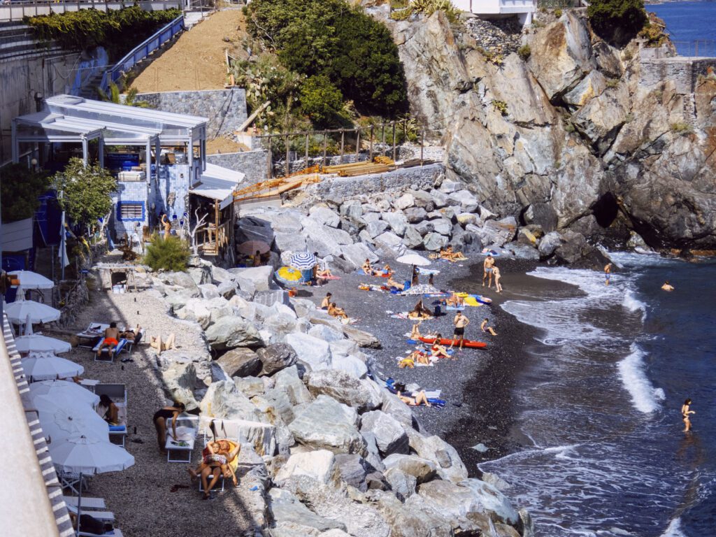 A monochrome photograph capturing a rocky beach scene. Sun loungers and umbrellas are scattered on the pebbled shore, with a few individuals lounging or walking around. The gentle surf breaks on the dark pebbles. To the left, a cliffside cafÃ© with an awning overlooks the beach, nestled between lush greenery and the rugged, steep rocks that form the backdrop. A concrete stairway is partially visible, descending towards the cafÃ© from above.