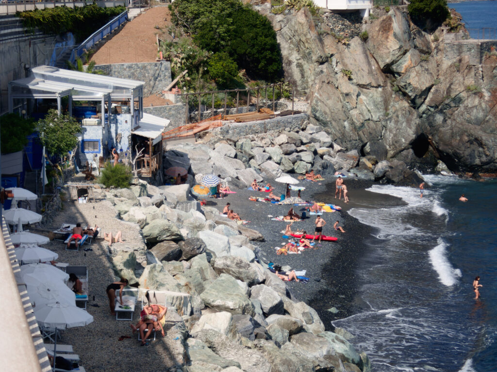 A photograph capturing a rocky beach scene. Sun loungers and umbrellas are scattered on the pebbled shore, with a few individuals lounging or walking around. The gentle surf breaks on the dark pebbles. To the left, a cliffside café with an awning overlooks the beach, nestled between lush greenery and the rugged, steep rocks that form the backdrop. A concrete stairway is partially visible, descending towards the café from above.