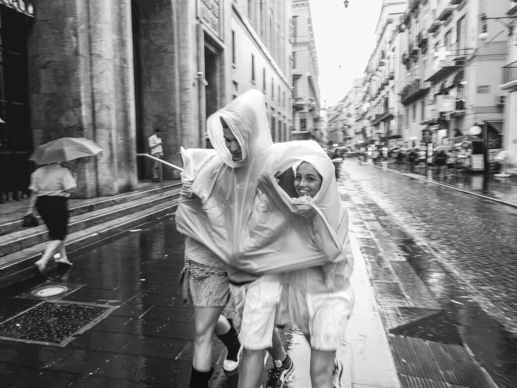 Three people, one with a visible joyful expression, share a light blue poncho in the rain on a city street. They seem to be in motion, with the surrounding environment slightly blurred, conveying a sense of bustling urban life despite the wet conditions. Other individuals with umbrellas are also captured in the scene, walking on the rain-slicked pavement that reflects the city lights.