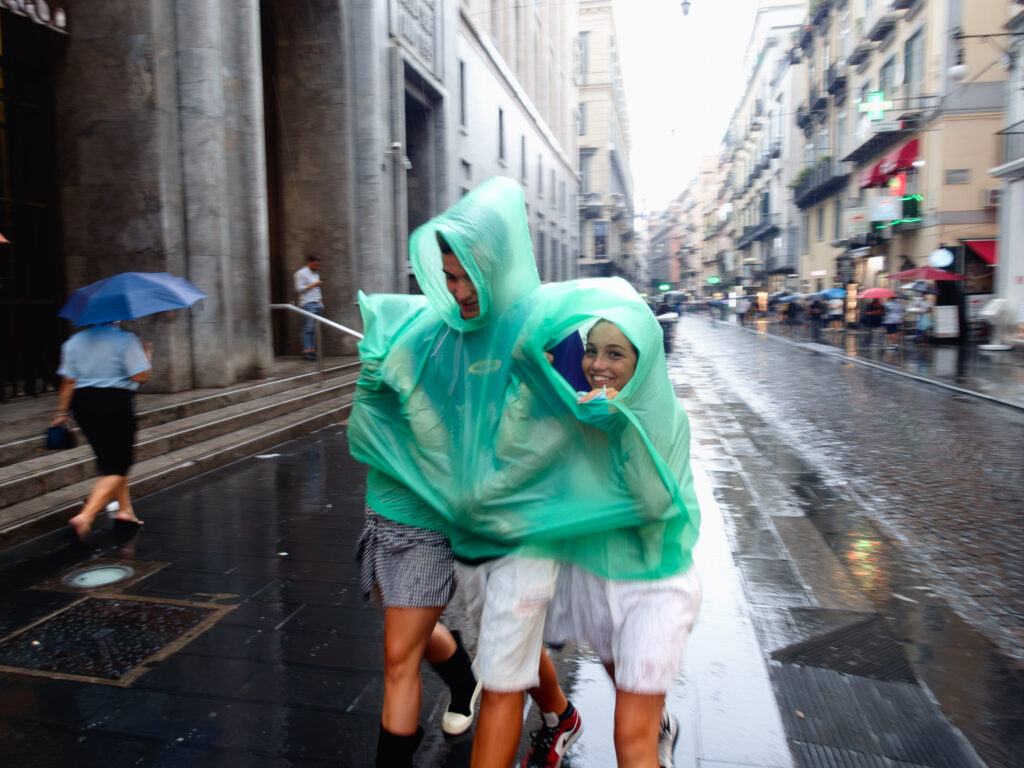 Three people, one with a visible joyful expression, share a light blue poncho in the rain on a city street. They seem to be in motion, with the surrounding environment slightly blurred, conveying a sense of bustling urban life despite the wet conditions. Other individuals with umbrellas are also captured in the scene, walking on the rain-slicked pavement that reflects the city lights.
