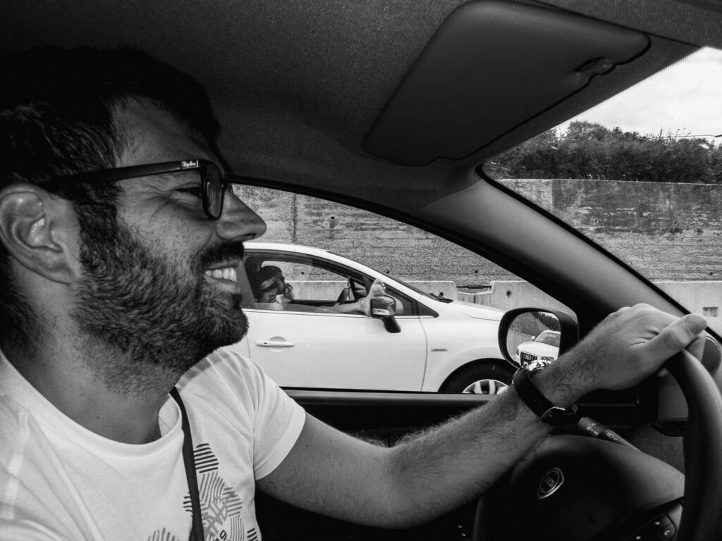 Black and white photo of a smiling man driving a car, wearing glasses and a white t-shirt. In the car ahead a girl sits in the passenger seat with her foot propped outside the window.