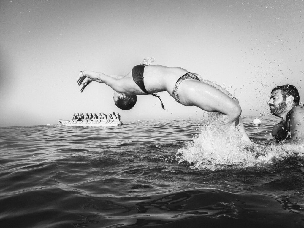 A black and white photograph captures a dynamic moment of two people in the sea. On the left, a person is mid-dive, their body curved gracefully in the air above the water surface, with droplets of water glistening around them. On the right, another person is swimming vigorously, creating a splash with their arms submerged in water. In the background, a group of people can be seen sitting on a large inflatable boat, observing the scene.