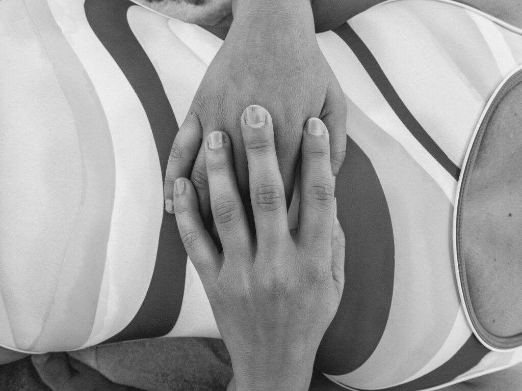 A close-up black and white photograph capturing the delicate placement of a hand over a striped pattern. The stripes curve and flow, likely part of a fabric or cushion, creating an abstract and artistic backdrop to the gently resting hand with unpolished nails, which adds a human element to the composition. The image plays with texture and form in a minimalist fashion.