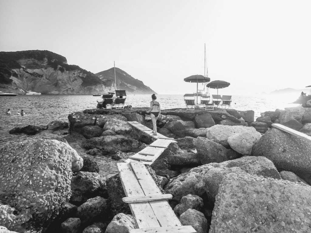 A black and white image of a rocky beachfront with a makeshift wooden walkway leading towards the water. A person stands midway on the plank path, looking towards the sea where a few people are swimming. There are lounge chairs with umbrellas set up on the rocks, suggesting a leisurely spot despite the rugged terrain. In the distance, a sailboat is anchored near the shore, with a hazy outline of mountains further back under a soft sky, adding a serene backdrop to this coastal scene.
