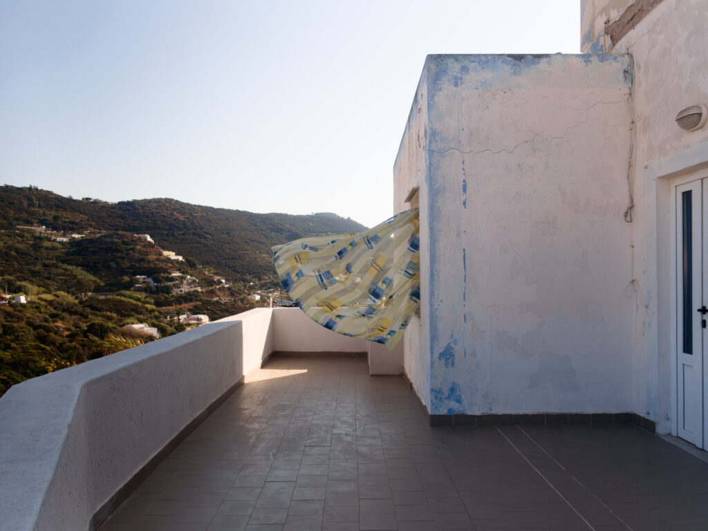 A terrace on a sunny day, with a large, billowing sheet caught in the wind creating dynamic folds in the air. The terrace is bordered by a low, smooth wall, and overlooks a hillside dotted with lush vegetation and scattered buildings. The wall of the building on the right shows signs of weathering and peeling paint, adding character to the scene. The contrast of the fluttering fabric and the sturdy architecture offers a sense of movement against the stillness of the landscape.