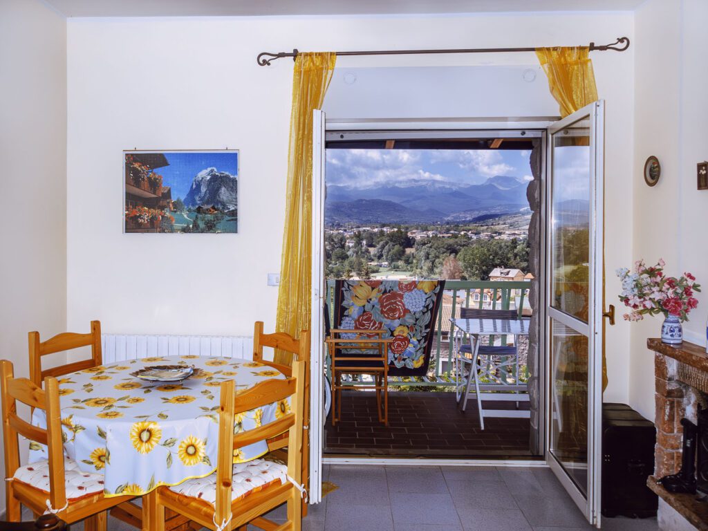 This is a black and white image of a cozy interior leading to a balcony with a picturesque view. Inside, there's a dining area with a round table covered with a floral tablecloth and surrounded by wooden chairs. On the wall hangs a framed picture of a mountainous landscape, mirroring the actual mountain view visible through the open balcony door. The balcony itself features outdoor furniture, offering a spot to enjoy the scenic backdrop of layered mountains stretching across the horizon, with a clear sky above and a glimpse of a small town nestled below.