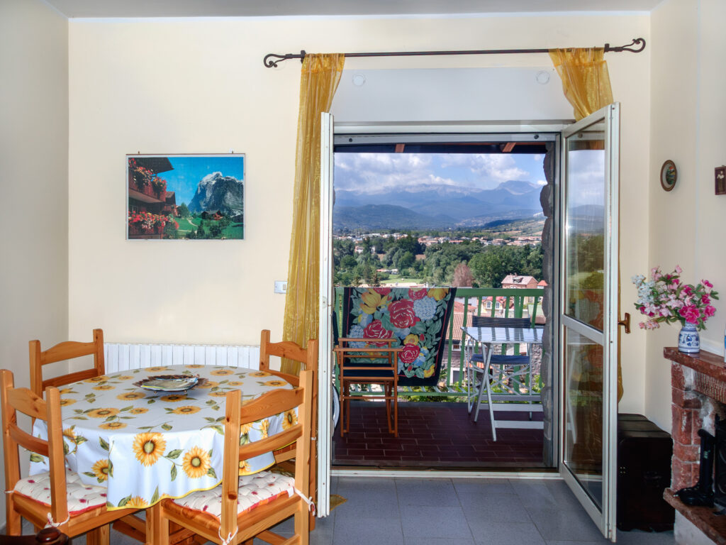 A cozy interior leading to a balcony with a picturesque view. Inside, there's a dining area with a round table covered with a floral tablecloth and surrounded by wooden chairs. On the wall hangs a framed picture of a mountainous landscape, mirroring the actual mountain view visible through the open balcony door. The balcony itself features outdoor furniture, offering a spot to enjoy the scenic backdrop of layered mountains stretching across the horizon, with a clear sky above and a glimpse of a small town nestled below.