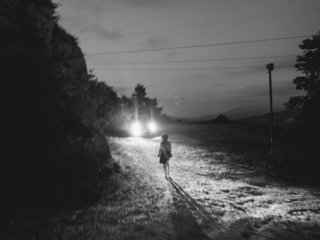 Photograph capturing a solitary figure walking on a rural road at night. The scene is illuminated by the headlights of an oncoming vehicle, casting a long shadow of the person on the rough, textured ground. The surroundings are dimly lit, with the outline of trees and a utility pole visible against the night sky, creating a moody and atmospheric shot.