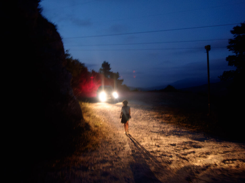 Photograph capturing a solitary figure walking on a rural road at night. The scene is illuminated by the headlights of an oncoming vehicle, casting a long shadow of the person on the rough, textured ground. The surroundings are dimly lit, with the outline of trees and a utility pole visible against the night sky, creating a moody and atmospheric shot.