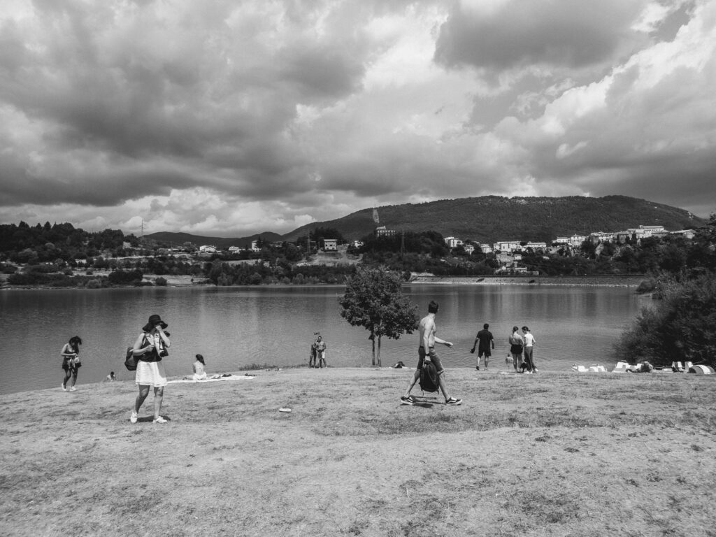 This is a black and white photograph of a relaxed, outdoor scene at a lakeside. People are scattered across the grassy shore; some are walking, while others are seated or standing by the water's edge. A woman in the foreground is wearing a wide-brimmed hat and appears to be taking a photo. On the left, an individual is walking away from the camera towards the water, and a few others are seen in the distance by the shoreline, possibly enjoying the view or preparing for a swim. The lake reflects the cloudy sky, and a backdrop of hills with buildings and trees completes the serene setting.