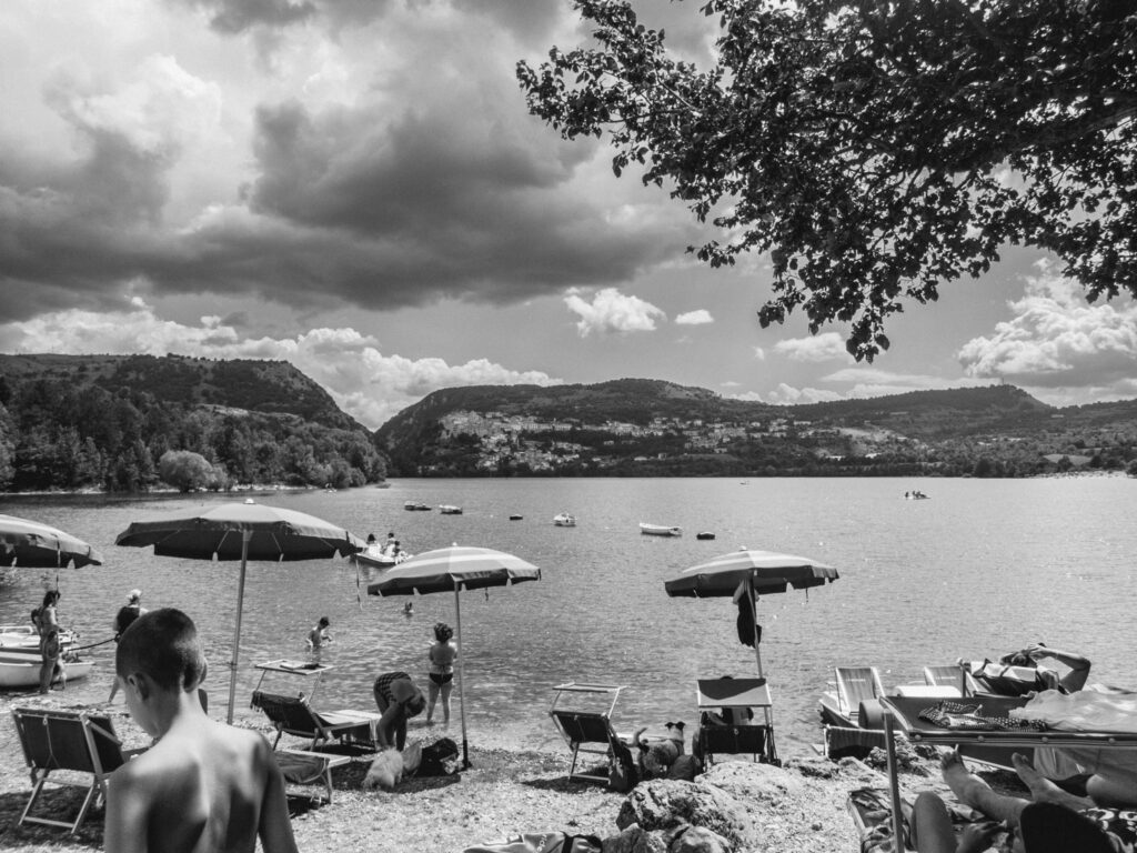 A monochromatic photograph captures a lively lakeside scene on a cloudy day. People relax on sun loungers under open umbrellas, while others partake in water activities like swimming and paddling small boats. The lake is serene, reflecting the gentle ripples of water activity. In the background, rolling hills dotted with trees and buildings rise from the opposite shore, completing a picturesque landscape. The presence of both leisure and nature creates a tranquil yet active atmosphere.