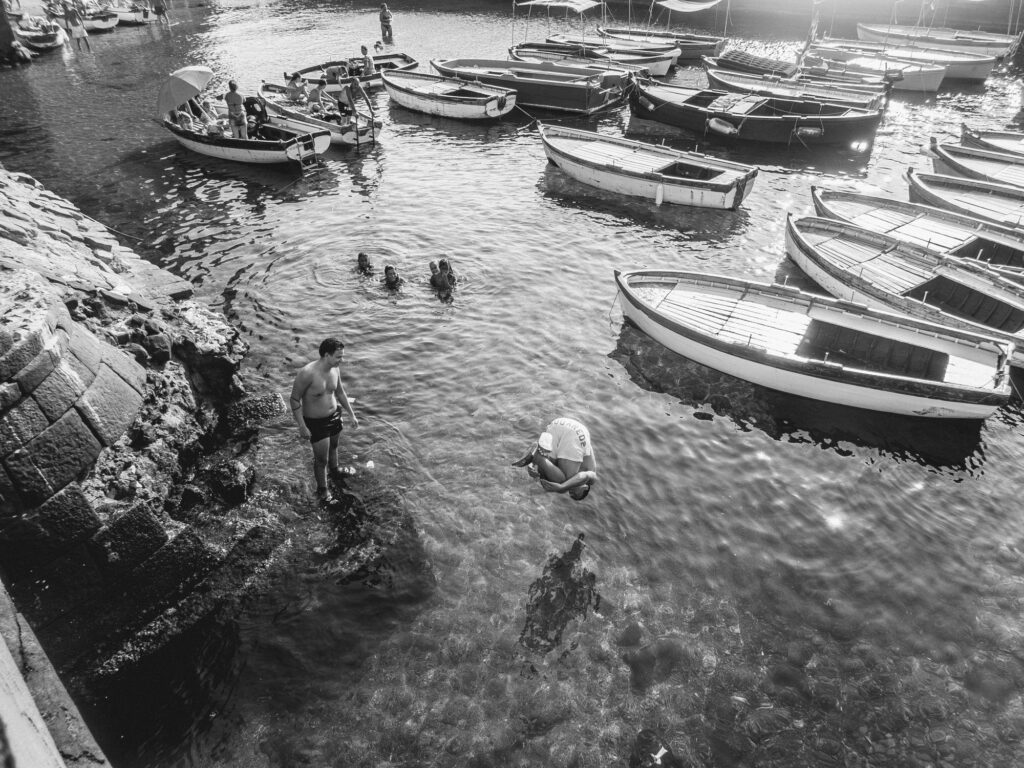 A monochrome seascape captures a moment of leisurely activity by the water: a cluster of small boats floats placidly in the sun-dappled sea while a group of people enjoys a swim. On the right, a young man in midair is seconds away from making a splash, adding a sense of imminent motion to the scene. Another individual, hunched over the rocks, seems engaged in an activity separate from the group. The scene is a harmonious blend of stillness and movement, framed by the rugged coastal rocks and the shimmering reflections on the water's surface.
