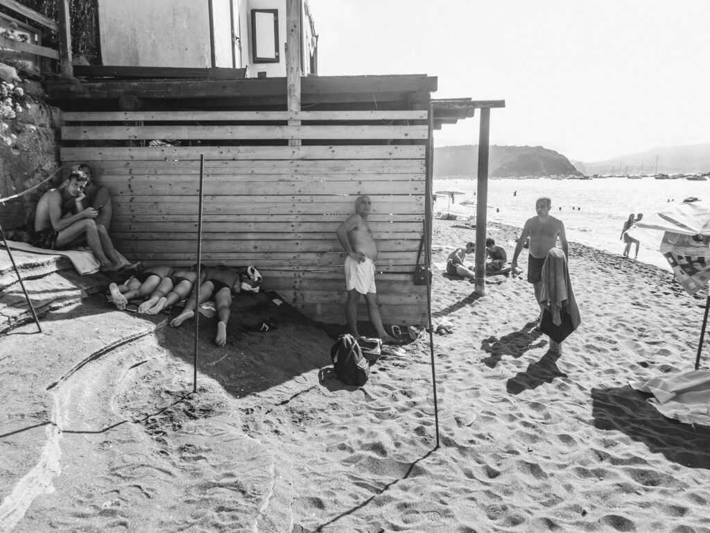Beach scene with several people engaging in typical seaside activities. On the left, a couple leans against a wooden shack, seeking shade, while two individuals lie on the sand, partially covered by a towel, napping. A man in bright green swim shorts stands in the center, resting his hand on his hip, looking off into the distance. To the right, another man walks across the sand holding a blue towel, with beachgoers sunbathing and swimming in the background. The atmosphere is relaxed with hills and boats visible across the water under the clear sky.